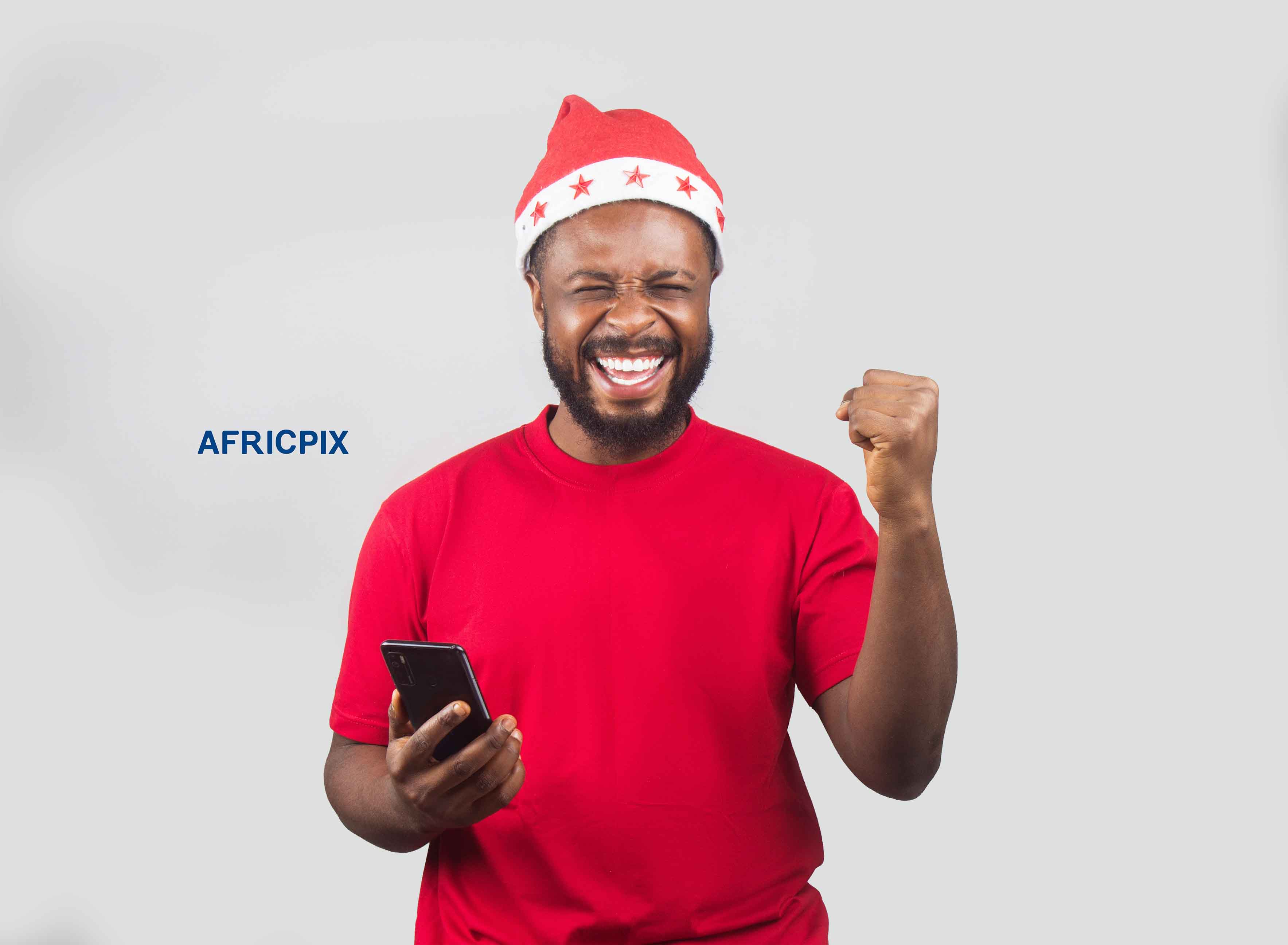 An African Nigerian man wearing a Christmas cap holding a phone, looking excited with his fist raised in celebration 1